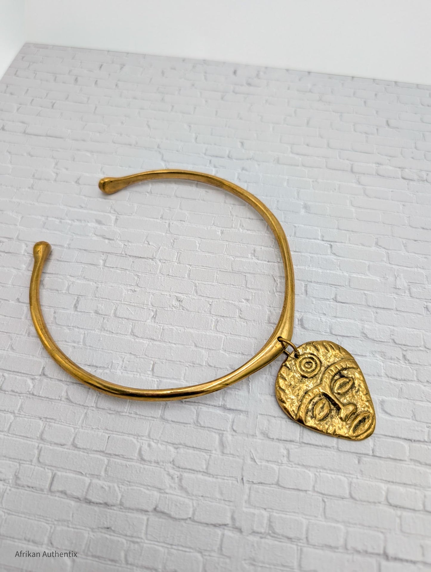 Gold collar necklace with an African mask pendant on a white brick background