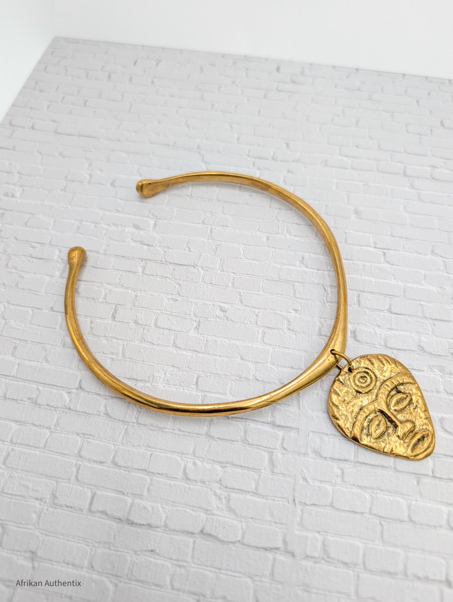 Gold collar necklace with an African mask pendant on a white brick background