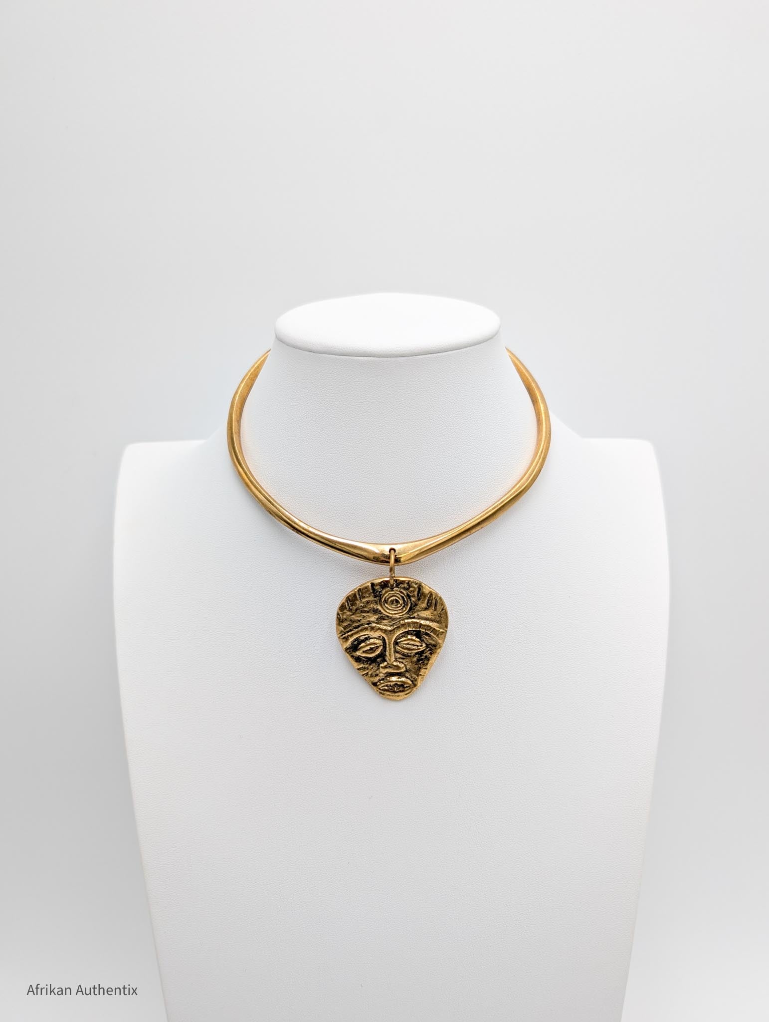 Gold collar necklace with an African mask pendant on a white mannequin bust against a gray background